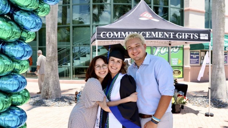 A mother graduates from college and hugs her children in front of balloons. She&#039;s wearing a cap and gown.