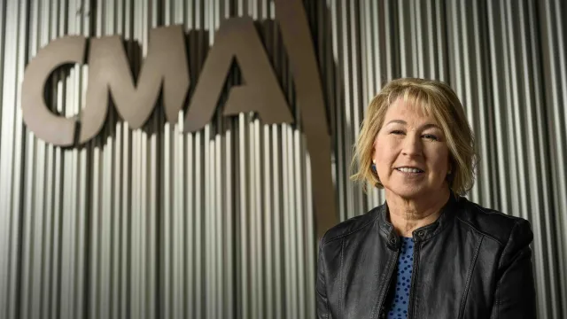 A white woman with blonde hair and a leather jacket smiles in front of a sign that says &quot;CMA.&quot;