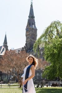 Alara Karahan graduation photo in a white dress with a blue stole with Healy Hall in the background