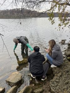 One male student wearing waders steps bends down into the Potomac River to collect water as another student and professor watch from the shoreline.