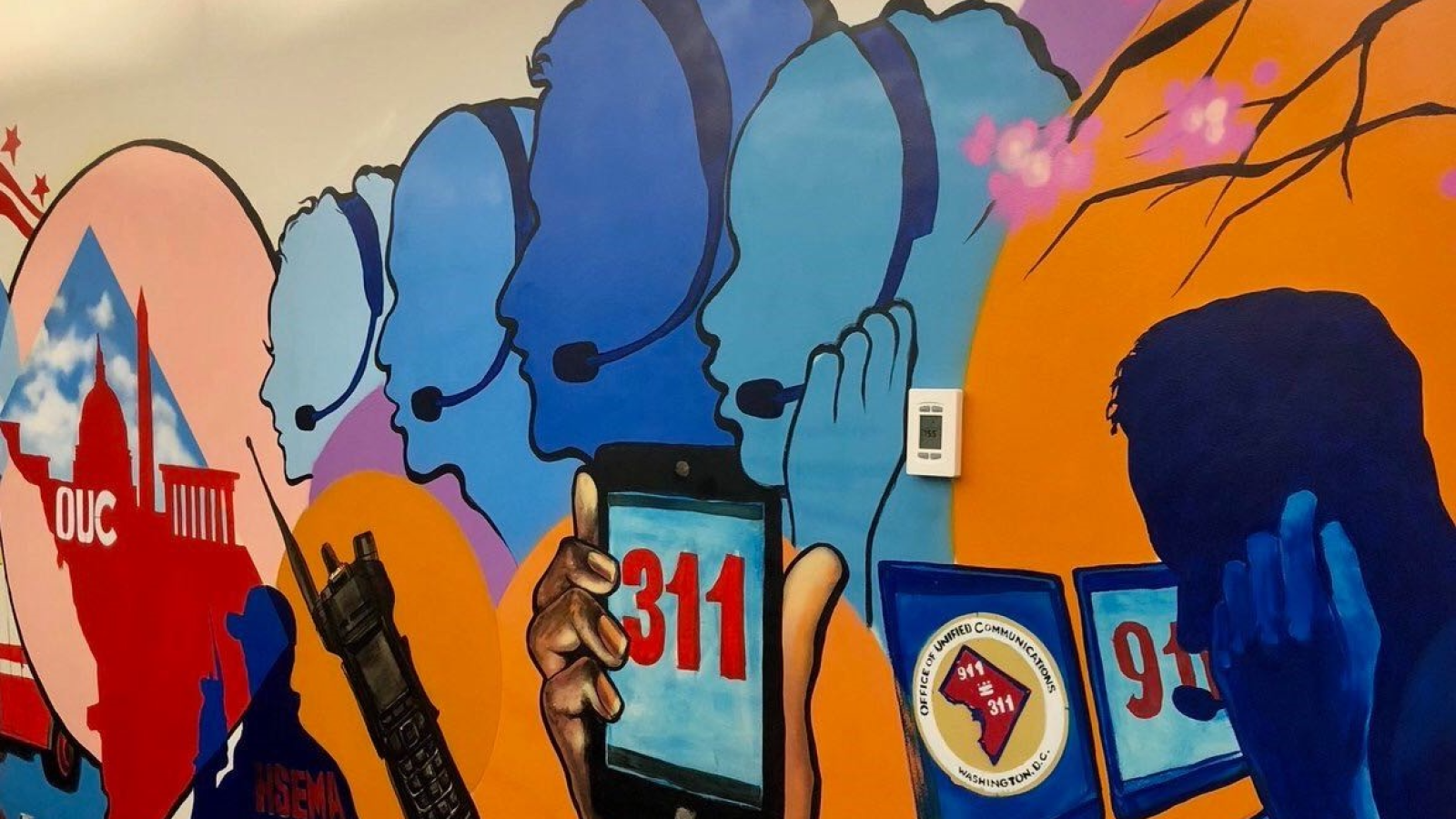A bright orange and blue mural of silhouettes of faces with a headset on and a hand holding up a phone that says &quot;311&quot; on the screen.