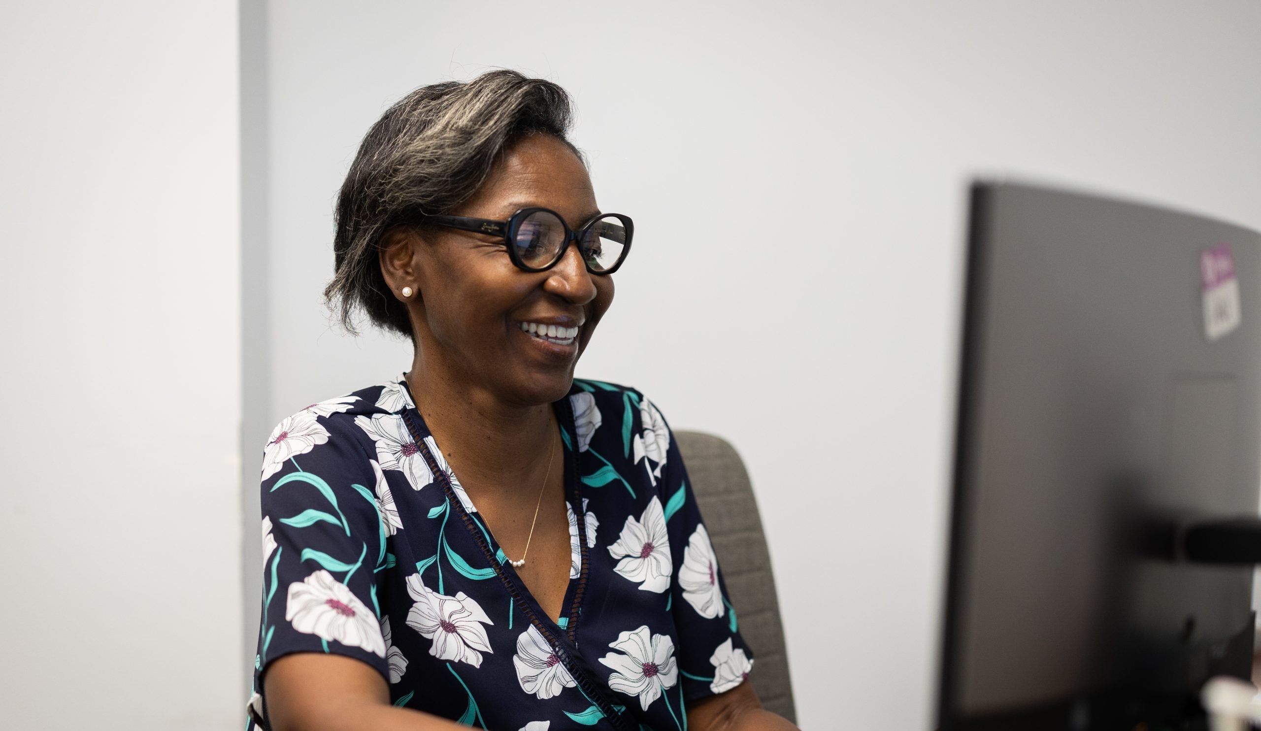 A woman with glasses and a black shirt with white flowers on it smiles as she looks at a computer.