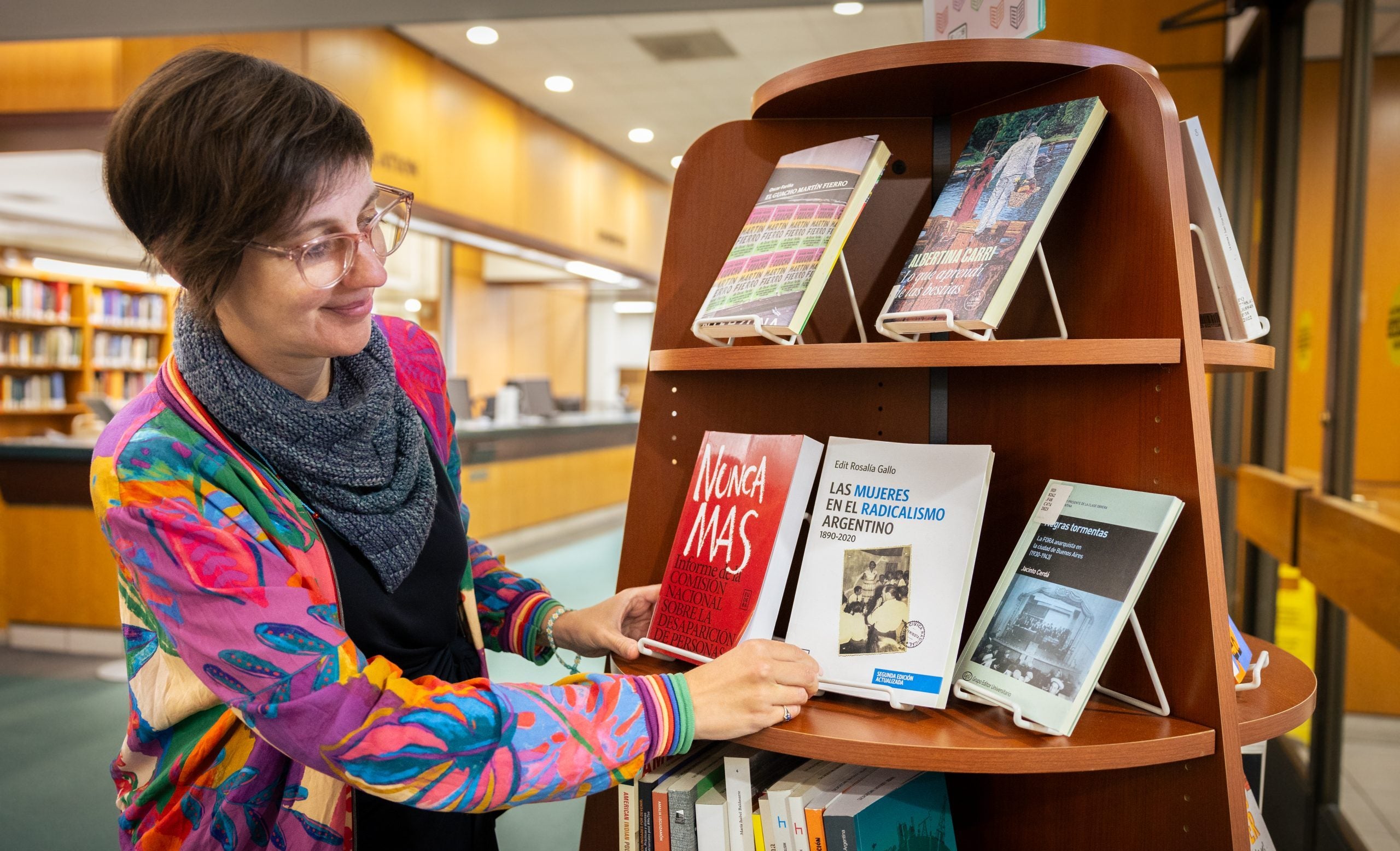 A woman in a colorful pink and blue jacket looks at a book on a shelf in a library.