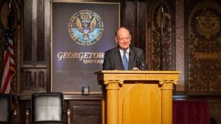 President DeGioia smiles behind a podium in Georgetown&#039;s Gaston Hall.