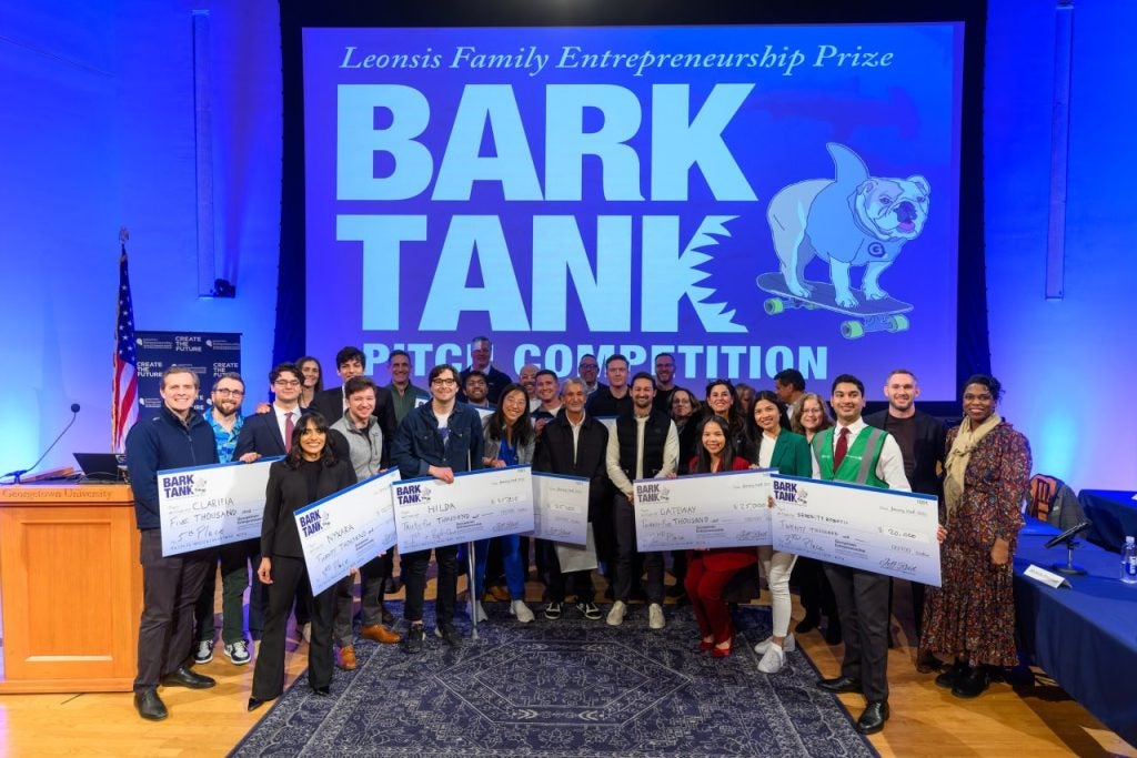 A group of students hold up giant checks in front of a blue screen that says "Bark Tank Pitch Competition"