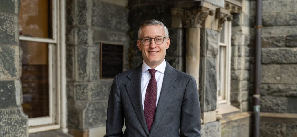 David Edelstein in a gray suit and dark red tie with hands in his pocket against Healy Hall background
