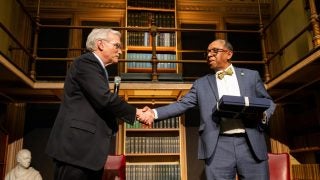 Two men shake hands on a stage with a bookshelf behind them.