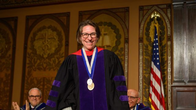 A woman in a purple academic robe smiles on a stage while wearing a medal