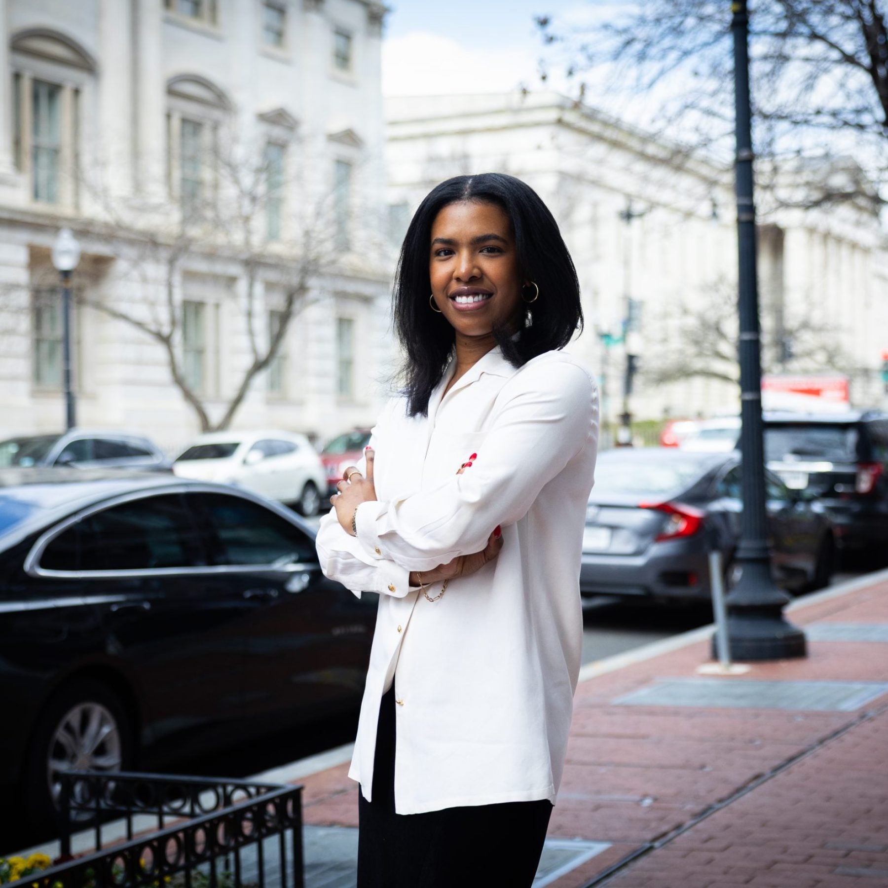 A woman stands with her arms crossed and smiles on the street in front of a white building