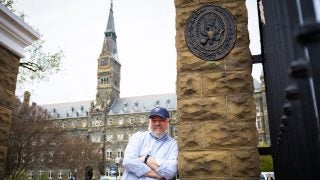 A man stands smiling with his arms crossed in front of a clock tower and university seal