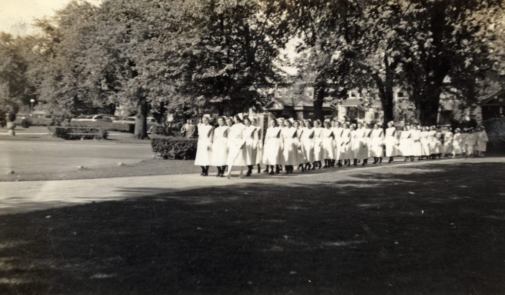 A group of nurses in white uniforms walk through campus in a black-and-white photo