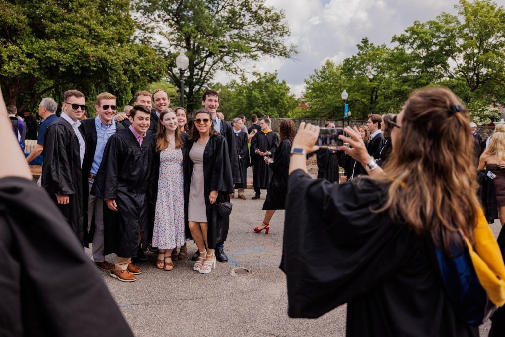 A group of graduates in caps and gowns pose for a group photo while someone else takes their photo on a phone.