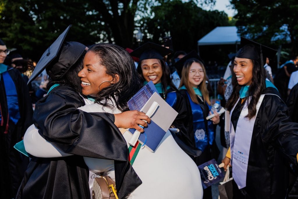 A Georgetown graduate in a cap and gown hugs another person while two other graduates look on.