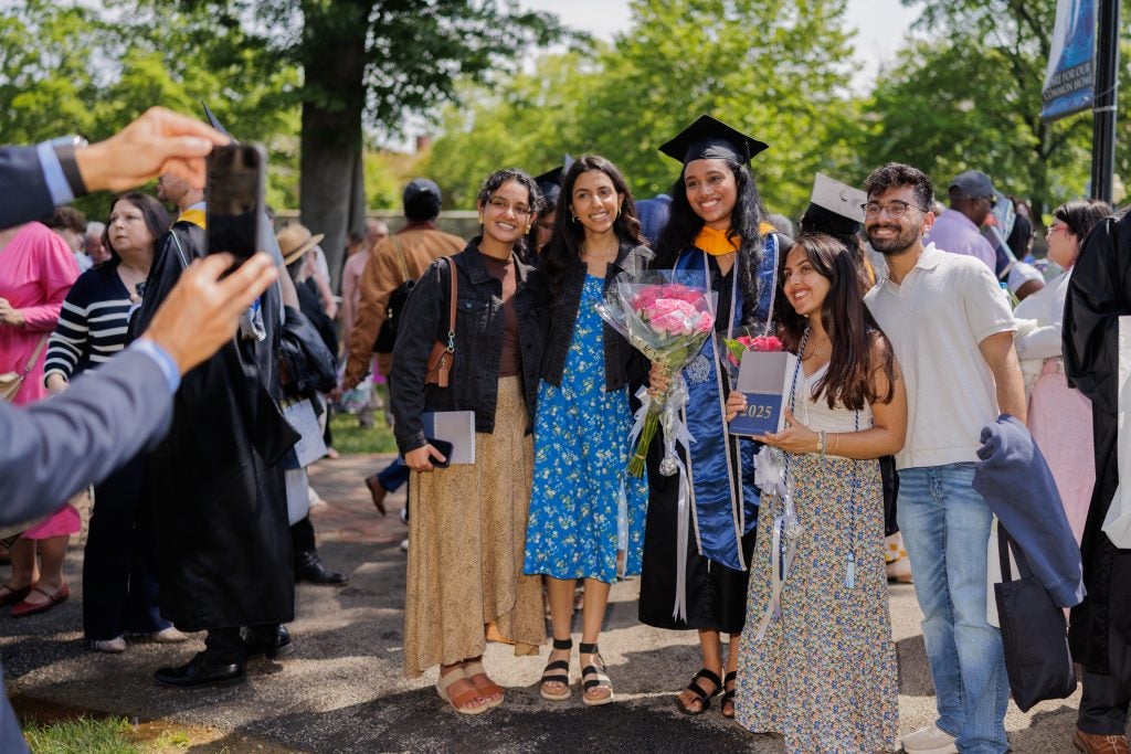 A graduate surrounded by three family members poses for a photo while someone takes that photo on a smartphone.