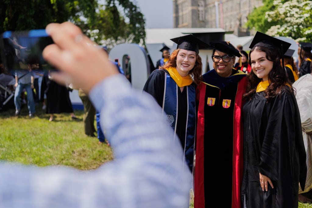 Two students in commencement regalia take a photo with a professor