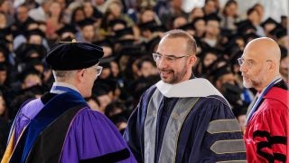 A priest in a graduation gown greets a faculty member onstage