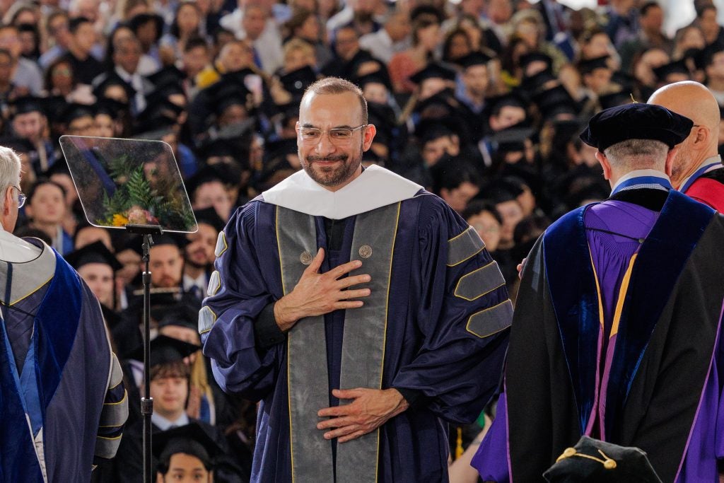 A priest in a purple graduation gown with a grey stole puts one hand over his heart