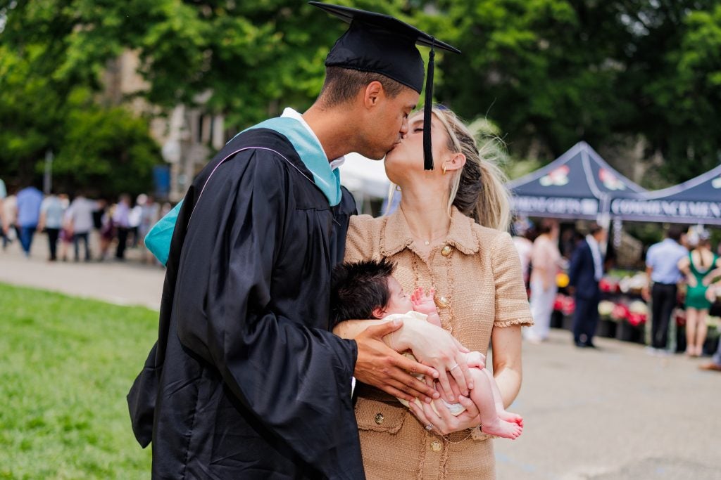 A young graduate kisses his wife while holding their baby