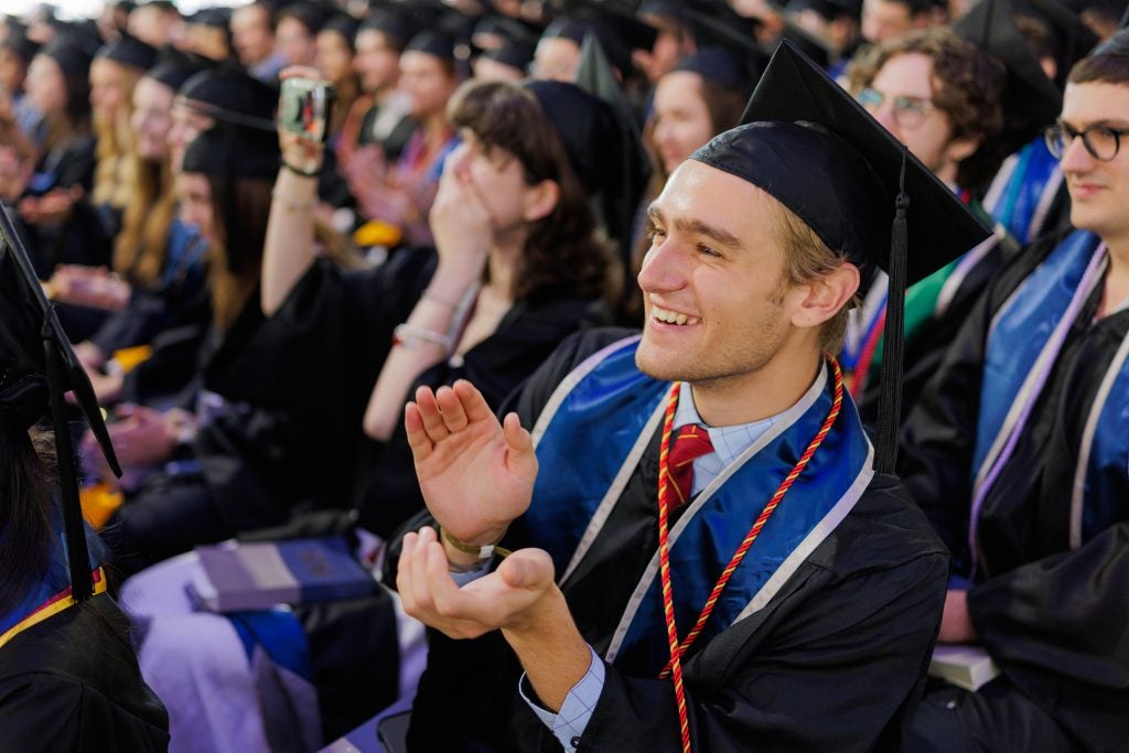 A graduate in a cap and gown sitting and clapping during a commencement ceremony.