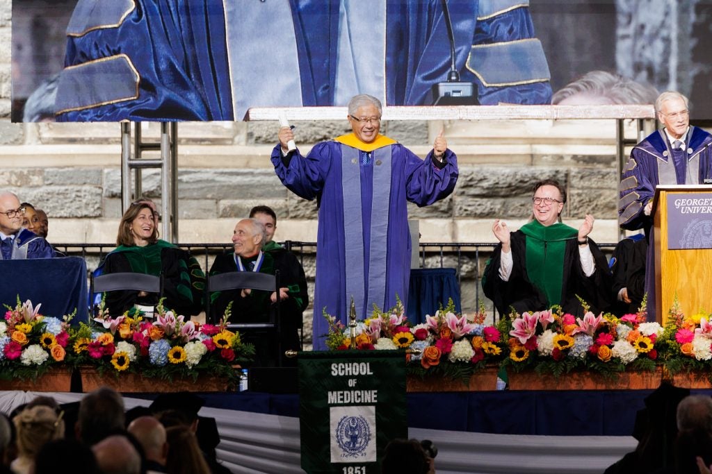 A man in a purple graduation gown holds up his arms onstage and smiles