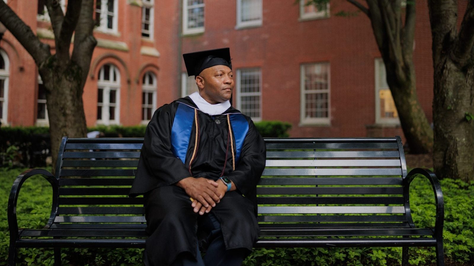 A man in a graduation gown and cap sits on a park bench