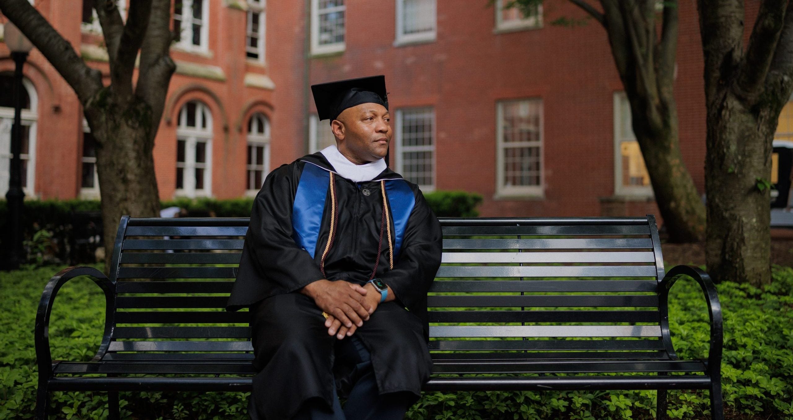 A man in a graduation gown and cap sits on a park bench