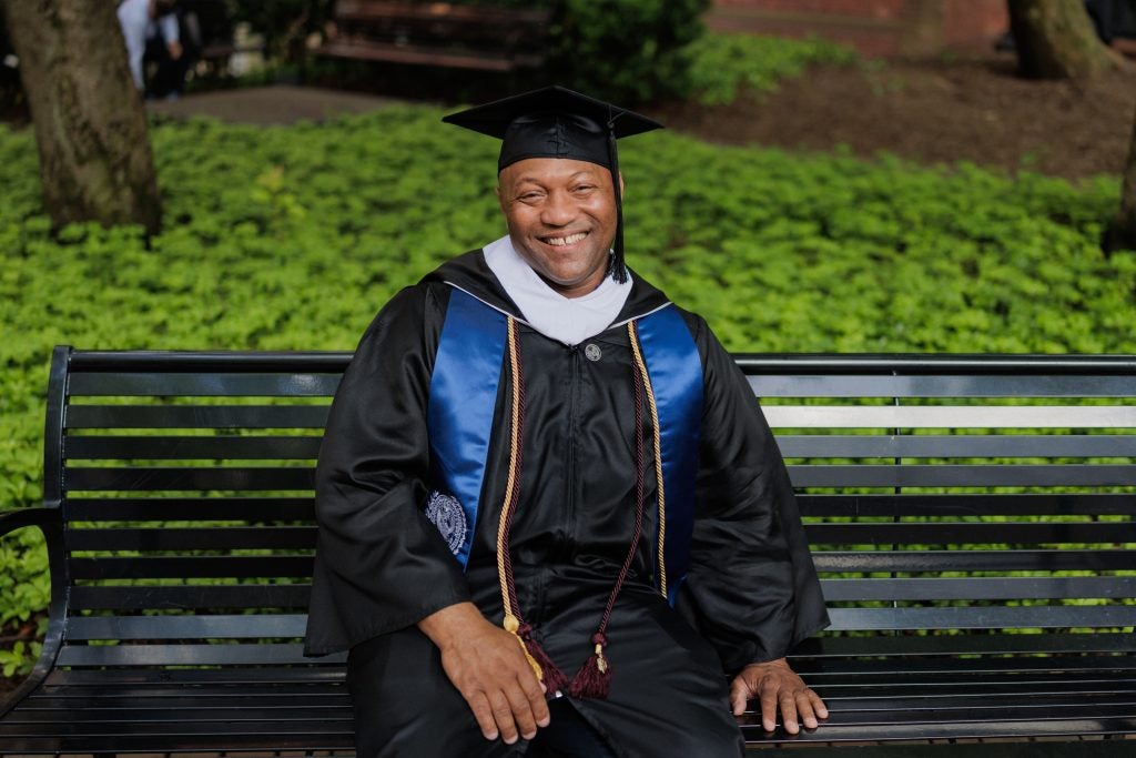 A man in a graduation cap and gown smiles on a park bench