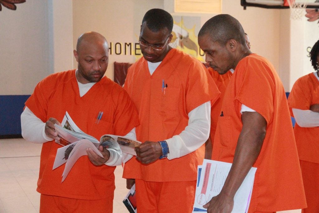 Three men in orange jumpsuits look at a book together inside a jail