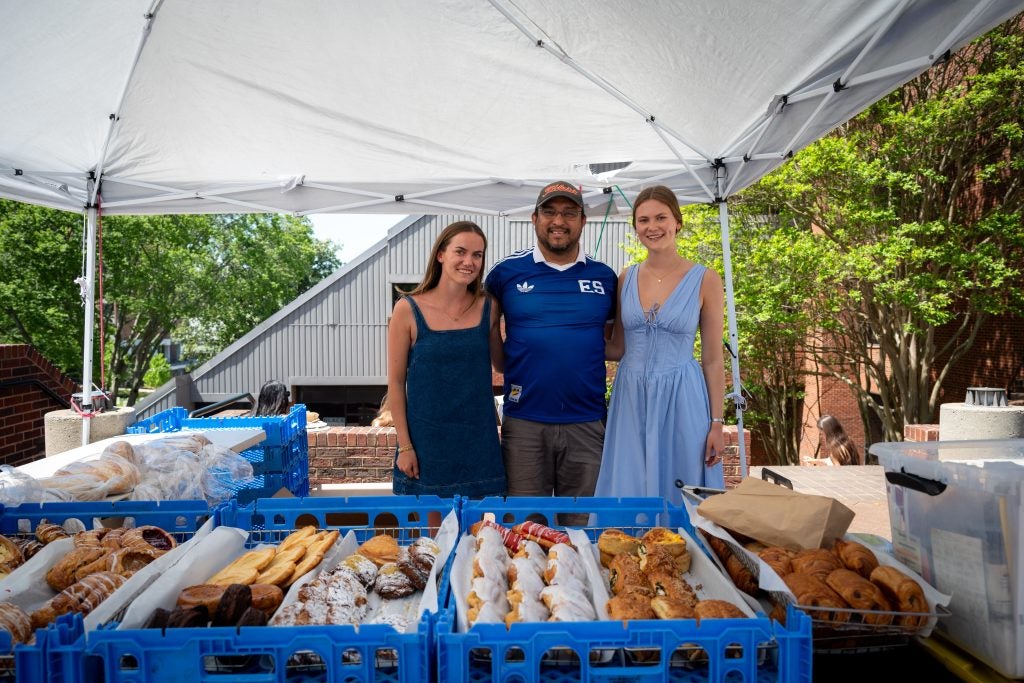 Two young women stand with a man in a blue shirt behind a stand full of pastries