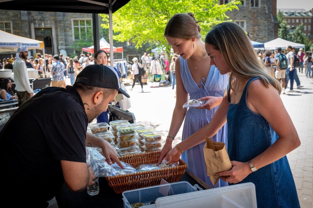 Two young women point at some food at a food stall with a staff member showing the food