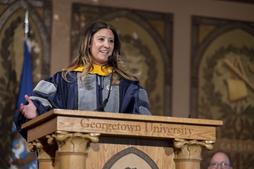 A woman in a purple and gray graduation gown speaks from behind a podium
