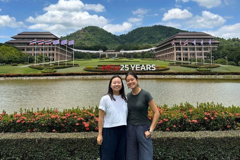 Two young Asian women outside with a body of water behind them and several buildings