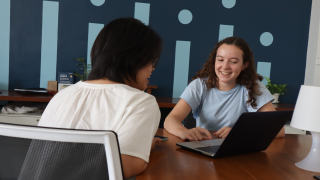 A young woman speaks to another woman while on a laptop