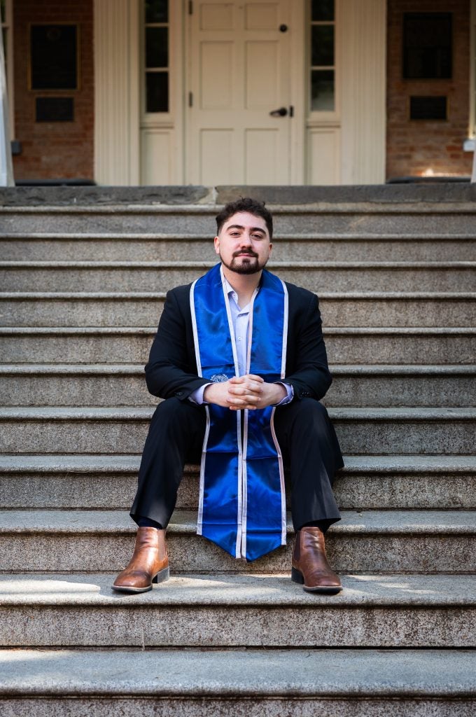 A young man wearing a blue graduation stole sits on stone steps with his fingers interlaced