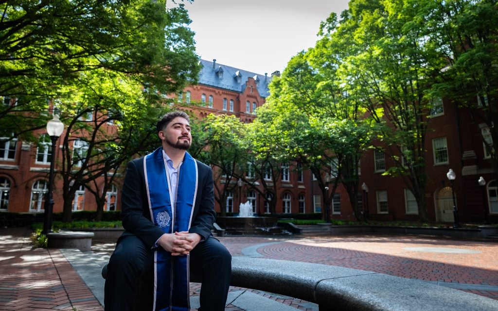 A young man wears a blue stole and sits with his fingers interlaced looking off into the distance. Behind him is a fountain