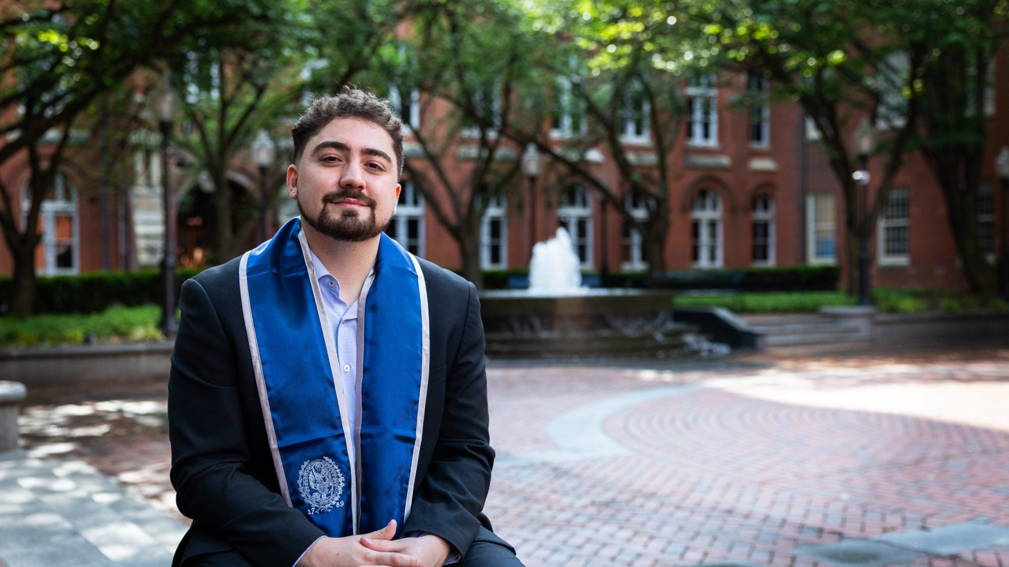 A young man wearing a blue graduation stole sits in a quad in front of a fountain