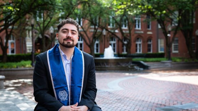 A young man wearing a blue graduation stole sits in a quad in front of a fountain
