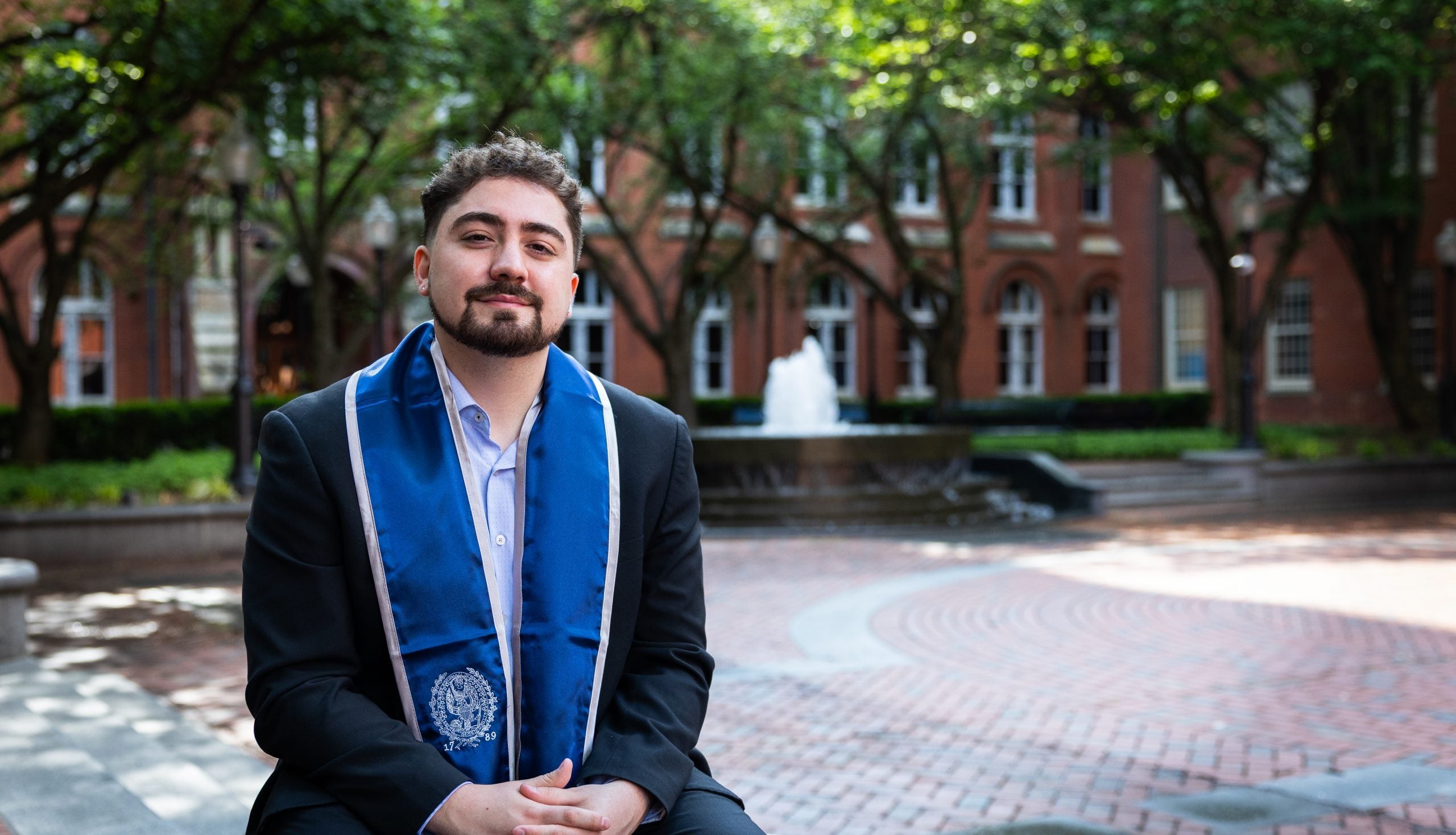 A young man wearing a blue graduation stole sits in a quad in front of a fountain