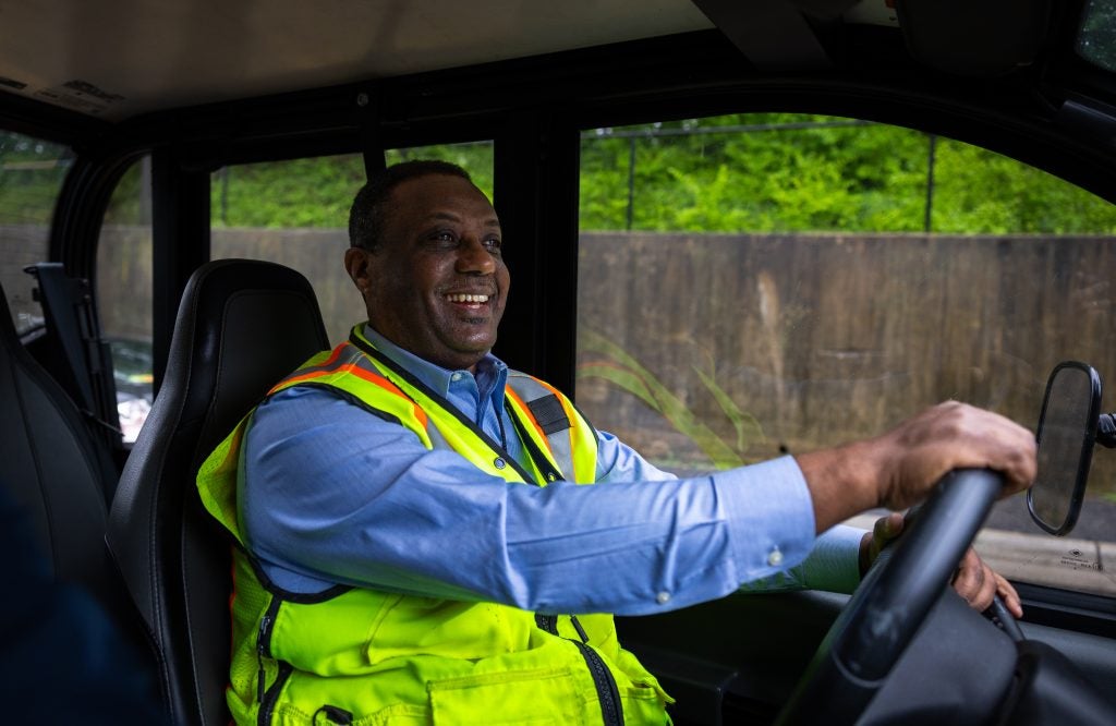A man in a yellow vest smiles as he drives a bus