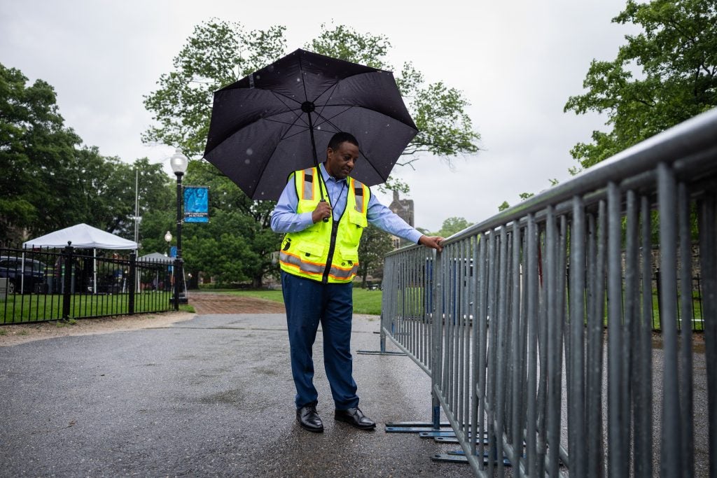 A man in a yellow vest holds an umbrella while straightening a gate used for graduation