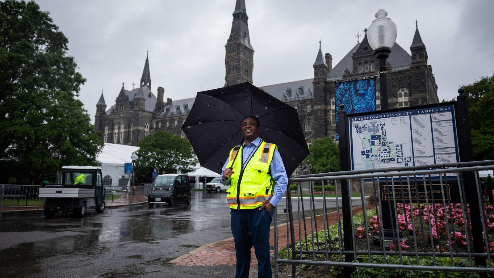 A man in a yellow vest holds an umbrella in front of Healy clocktower on Georgetown&#039;s campsu