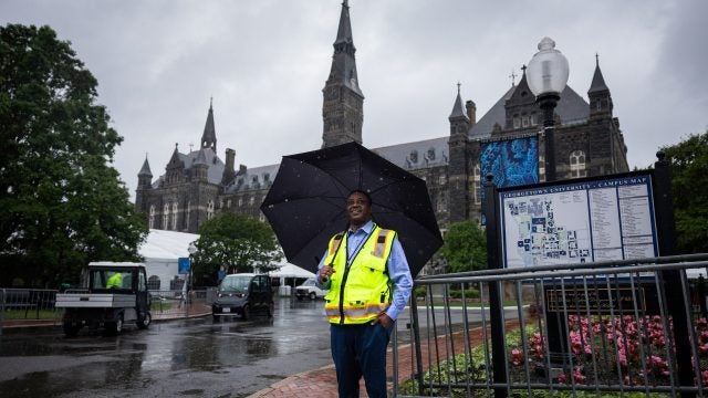 A man in a yellow vest holds an umbrella in front of Healy clocktower on Georgetown&#039;s campsu