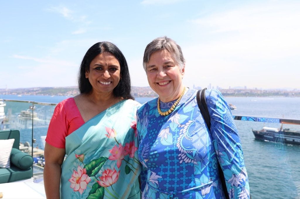 Two women smile on a rooftop overlooking the sea