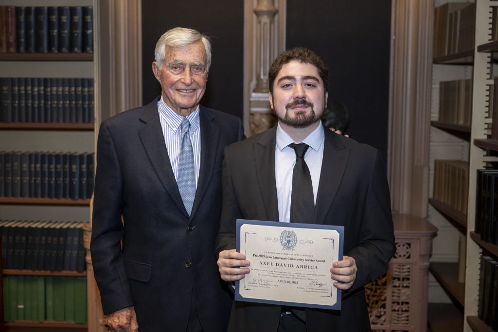 A young man holds an award with an older man. Both wear suits