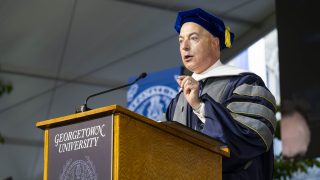 A man in a graduation gown stands behind a podium
