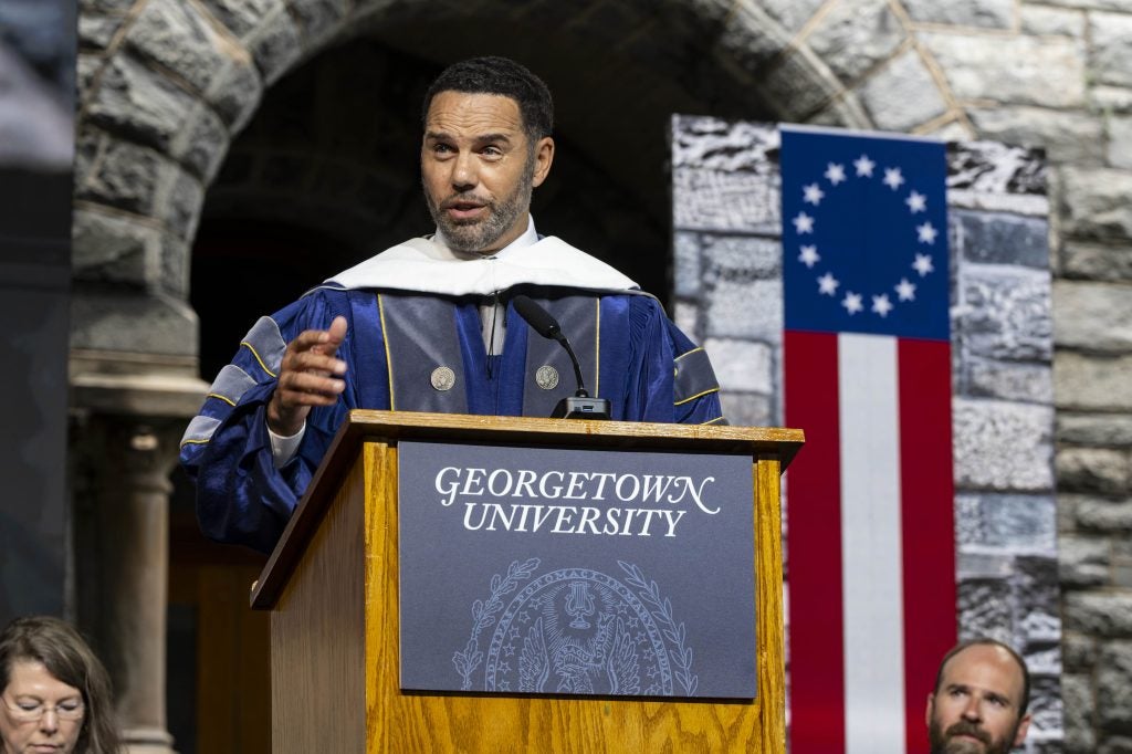 A man in a blue and purple graduation gown delivers a speech from behind a podium that says Georgetown University on it