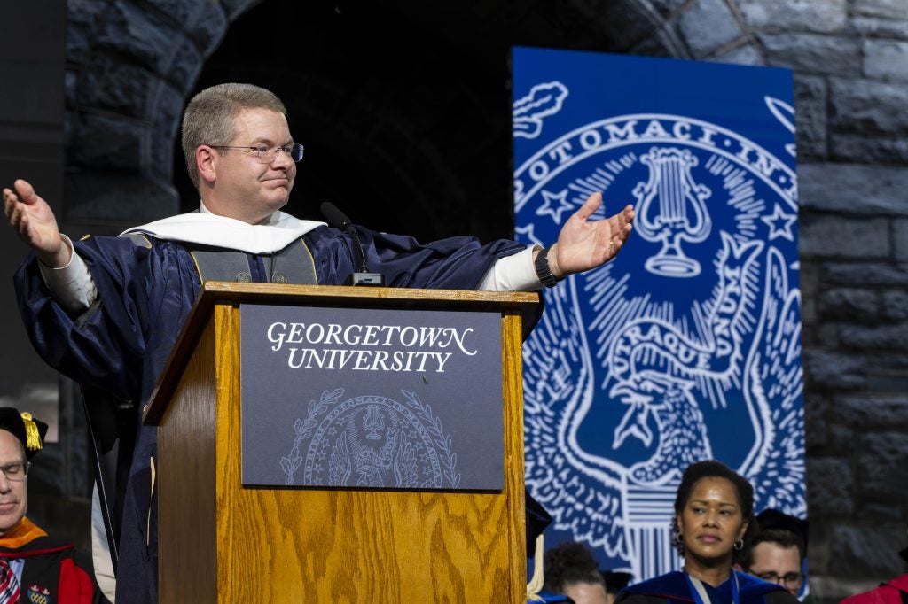 A man in a graduation gown holds out both his arms to a crowd from behind a podium