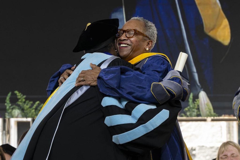 Two people in graduation gowns embrace onstage