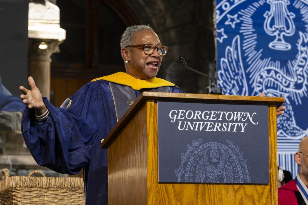 A woman wearing a purple graduation gown and a yellow stole speaks from behind a podium