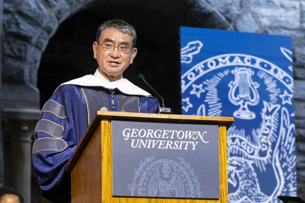 A man in a gray and purple graduation gown speaks behind a Georgetown podium